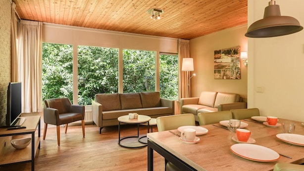 A cozy room with a wooden ceiling, a sofa, a dining table set for four, and a large window with a view of greenery. - Bungalow in Putten with COPD-Friendly Design (Putten)
