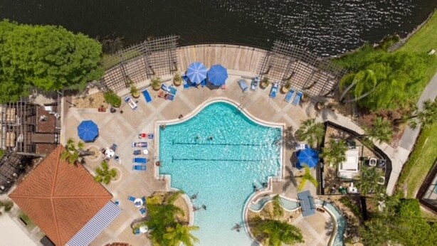 An aerial view of a resort with a central swimming pool, sun loungers, and palm trees. - Amazing 3BR 2BA Townhome With Lake View at Terra Verde! (Kissimmee)