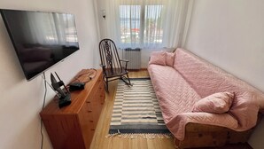 Een kleine, knusse kamer met een flatscreen televisie aan de muur, een houten bureau met een stoel, een roze gewatteerde bank en een gestreept vloerkleed.