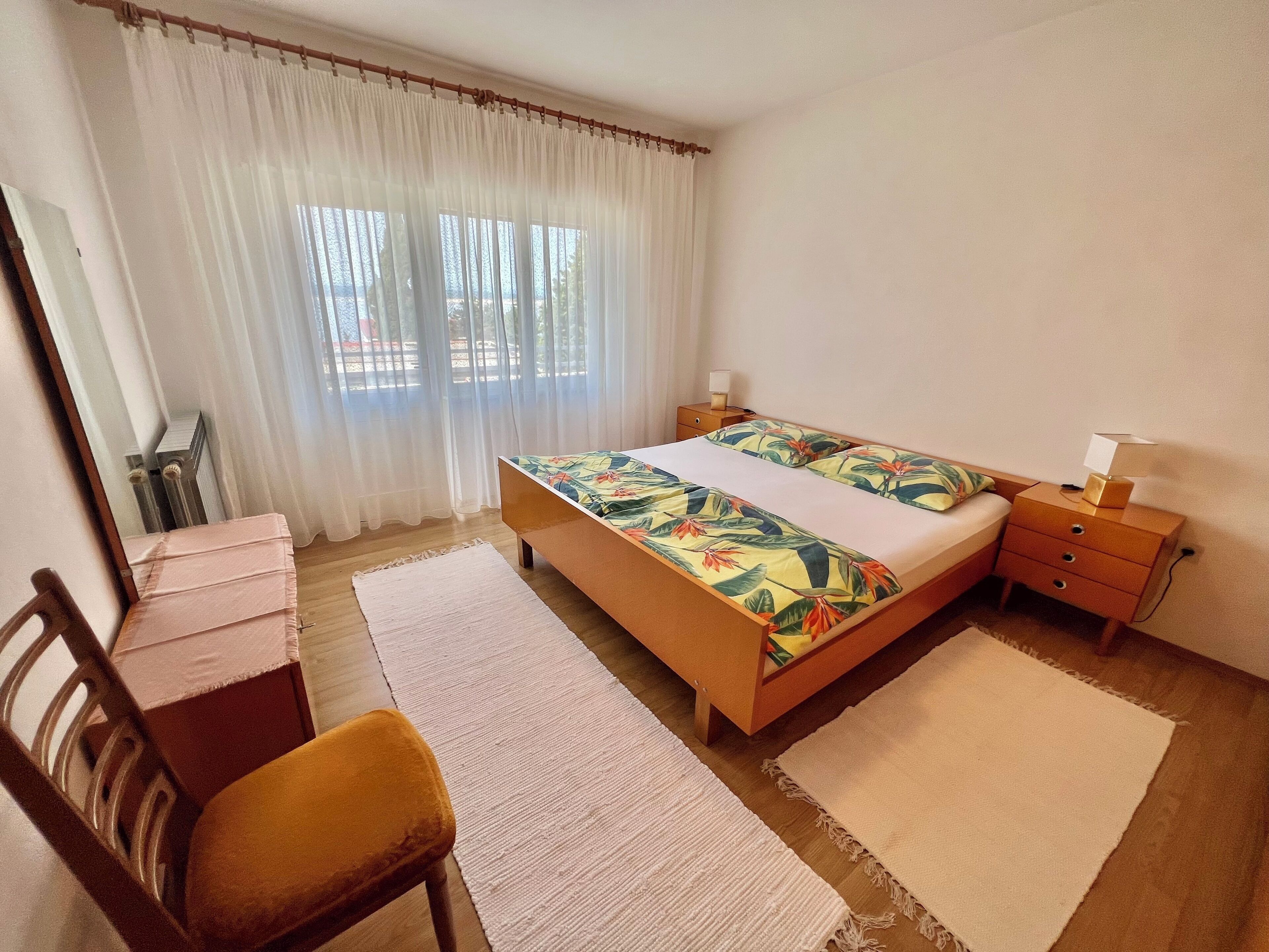 A bedroom with a bed, a nightstand, a chair, a desk, and a window with curtains.