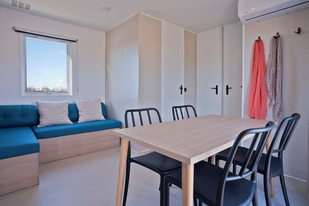 A modern room with a sofa, a dining table, and chairs. There is a window with blinds, a door with a handle, and a wall-mounted air conditioner.