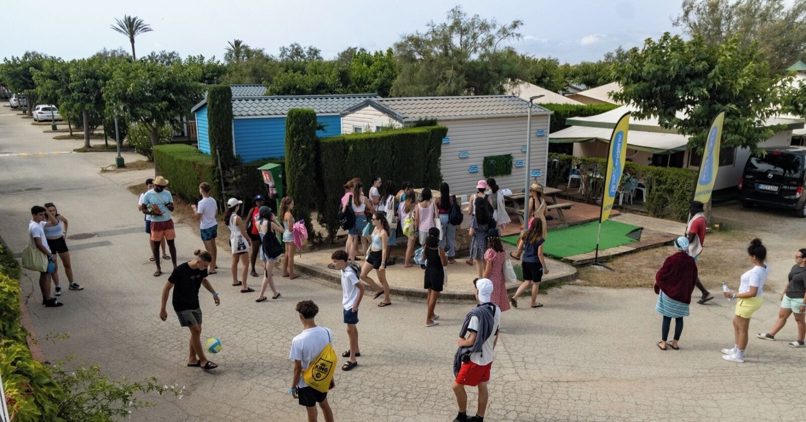 A group of people gathered in a courtyard with a small building, trees, and a parked car.