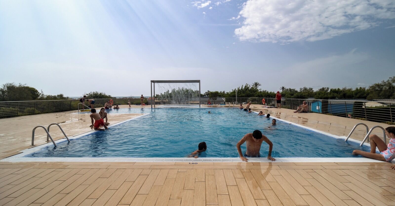 Una piscina con gente disfrutando del agua, rodeada por una terraza de madera y barandales de metal.