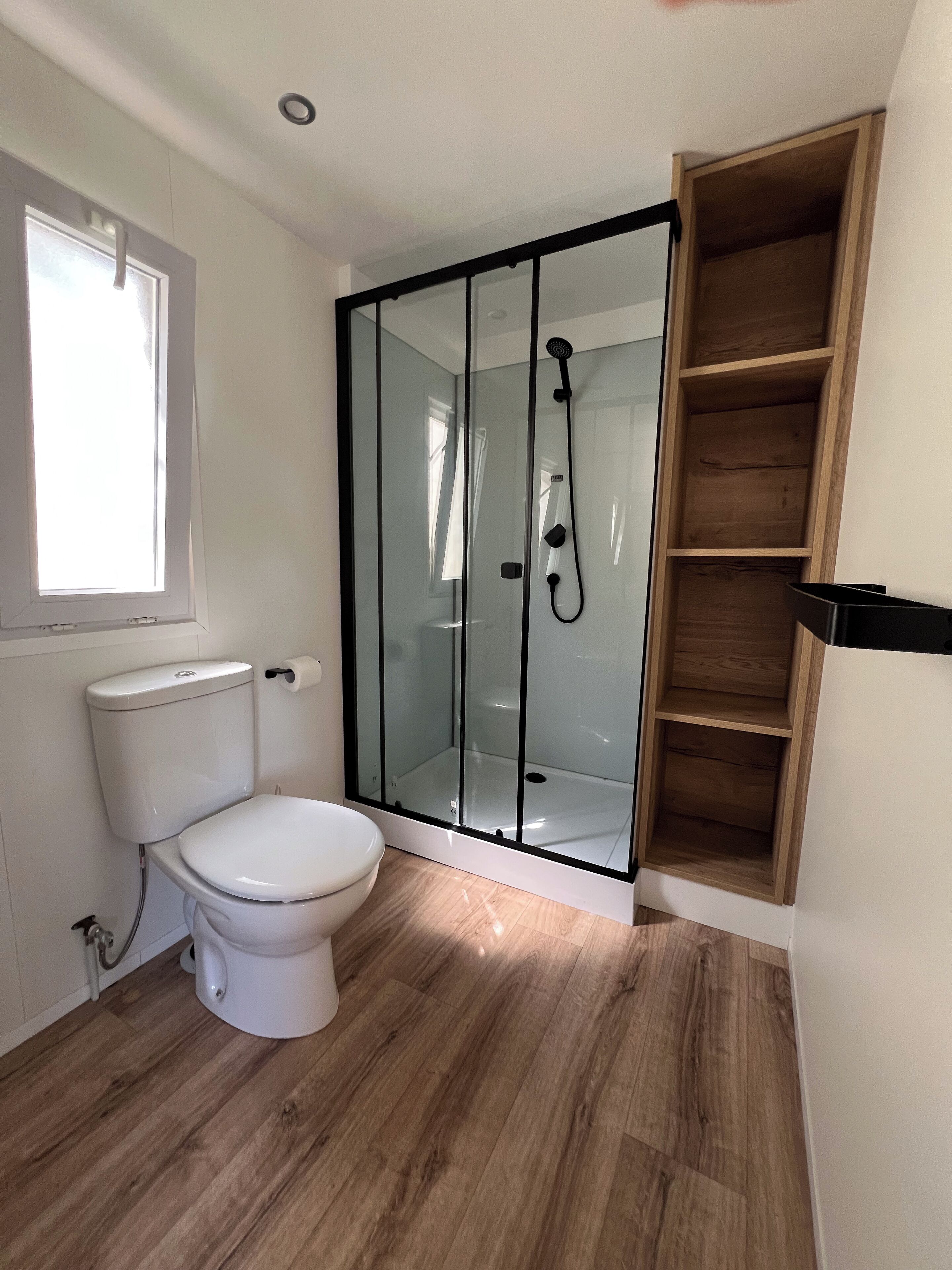 Un baño con inodoro, ducha con mampara de vidrio y un mueble de almacenamiento de madera.