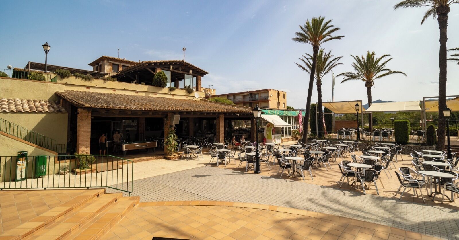 An outdoor café area with tables and chairs, a building with a tiled roof, and palm trees.