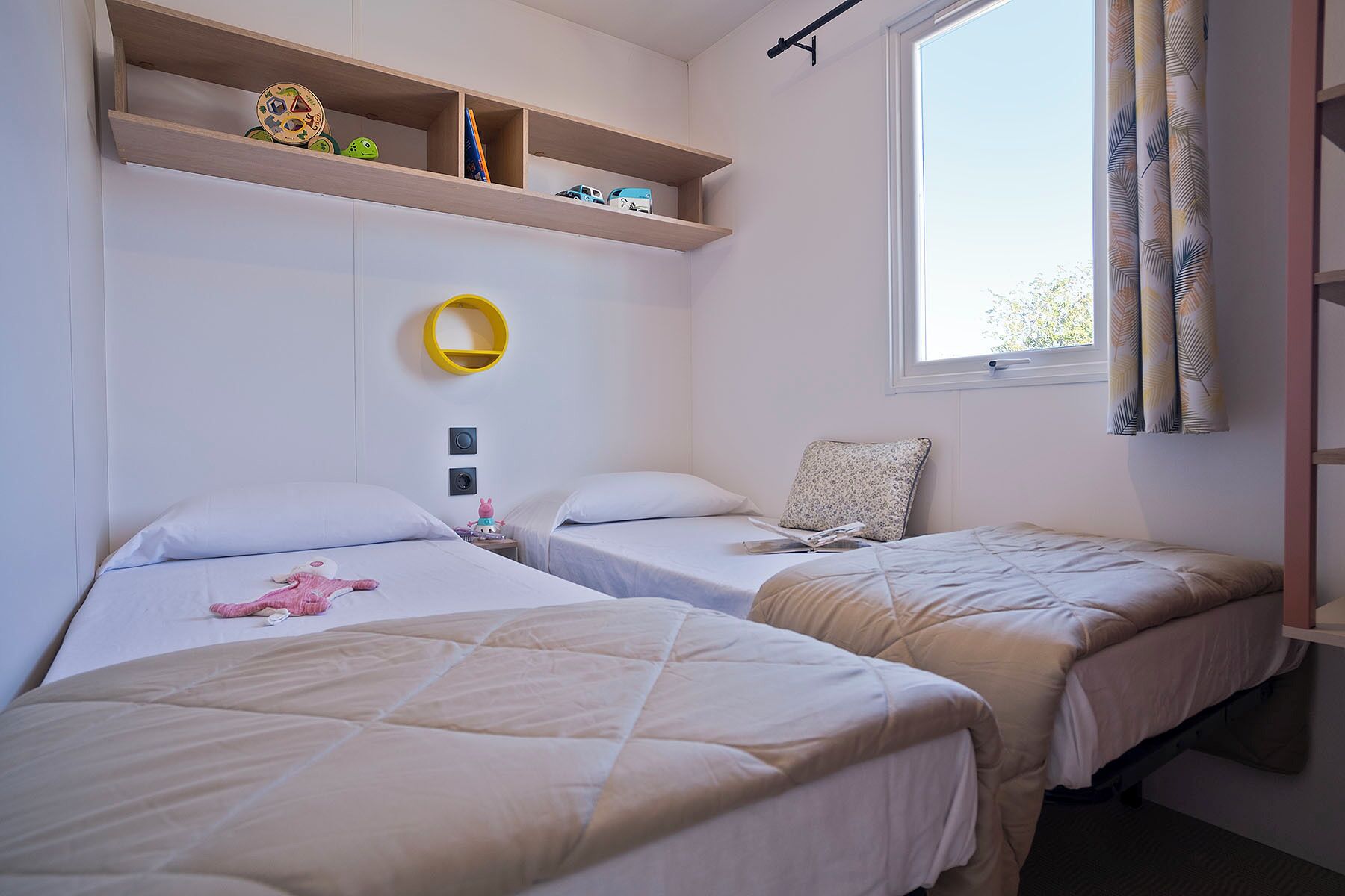 A room with two beds, a shelf with toys, a small table, and a window with curtains.