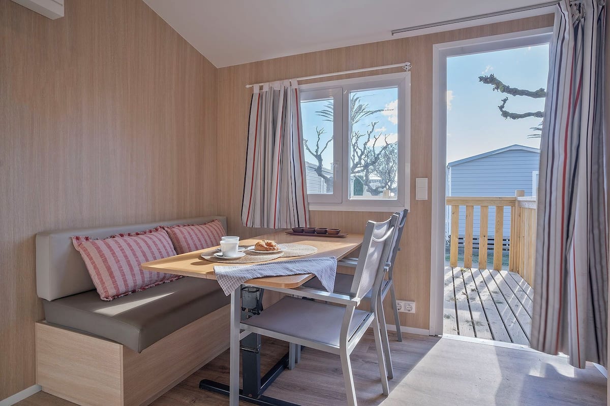 A cozy room with a wooden bench, a dining table, and a chair. There are two windows with curtains, and a view of a wooden deck outside.