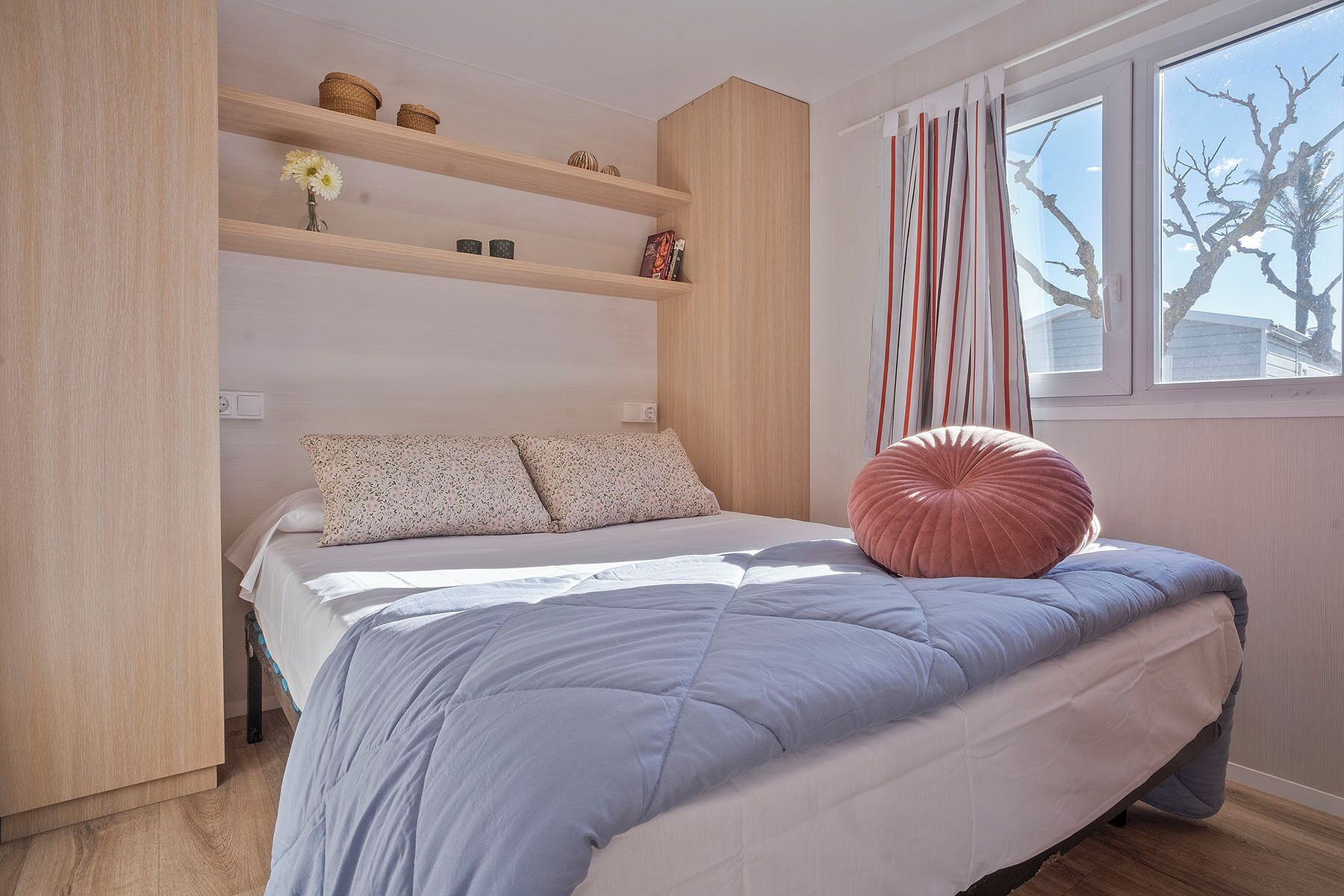 Une chambre à coucher avec un lit, une fenêtre avec des rideaux et une étagère ornée d’objets décoratifs.