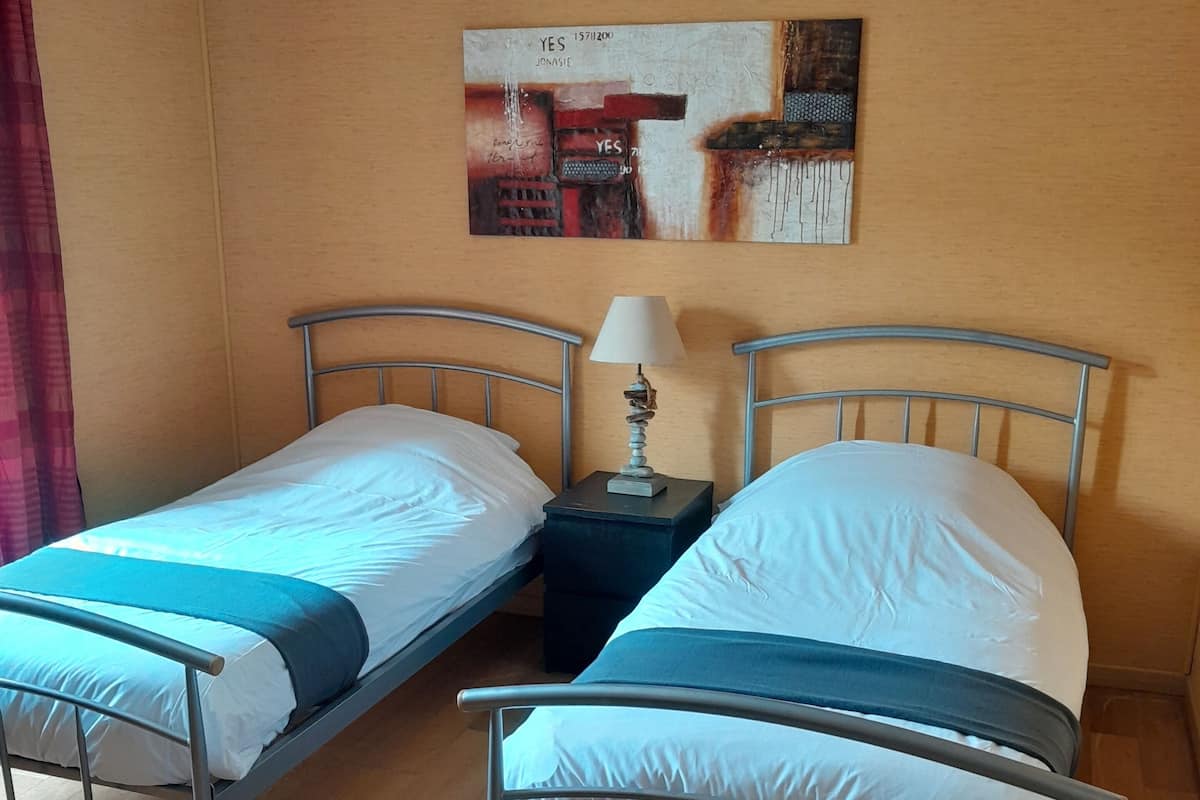 A room with two metal beds, a nightstand with a lamp, and a painting on the wall.