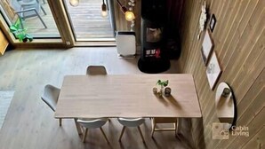 A dining area with a wooden table, two chairs, and a small plant. There is a window with a view of the outside, and a black speaker in the background. - Sjusjøen ski in/out, ski storage, Wifi, grill & EV-charger, modern apartment! (Sjusjøen)