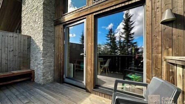 A wooden deck with a stone wall, a sliding glass door leading to an outdoor seating area, and a view of trees and sky. - Sjusjøen ski in/out, ski storage, Wifi, grill & EV-charger, modern apartment! (Sjusjøen)