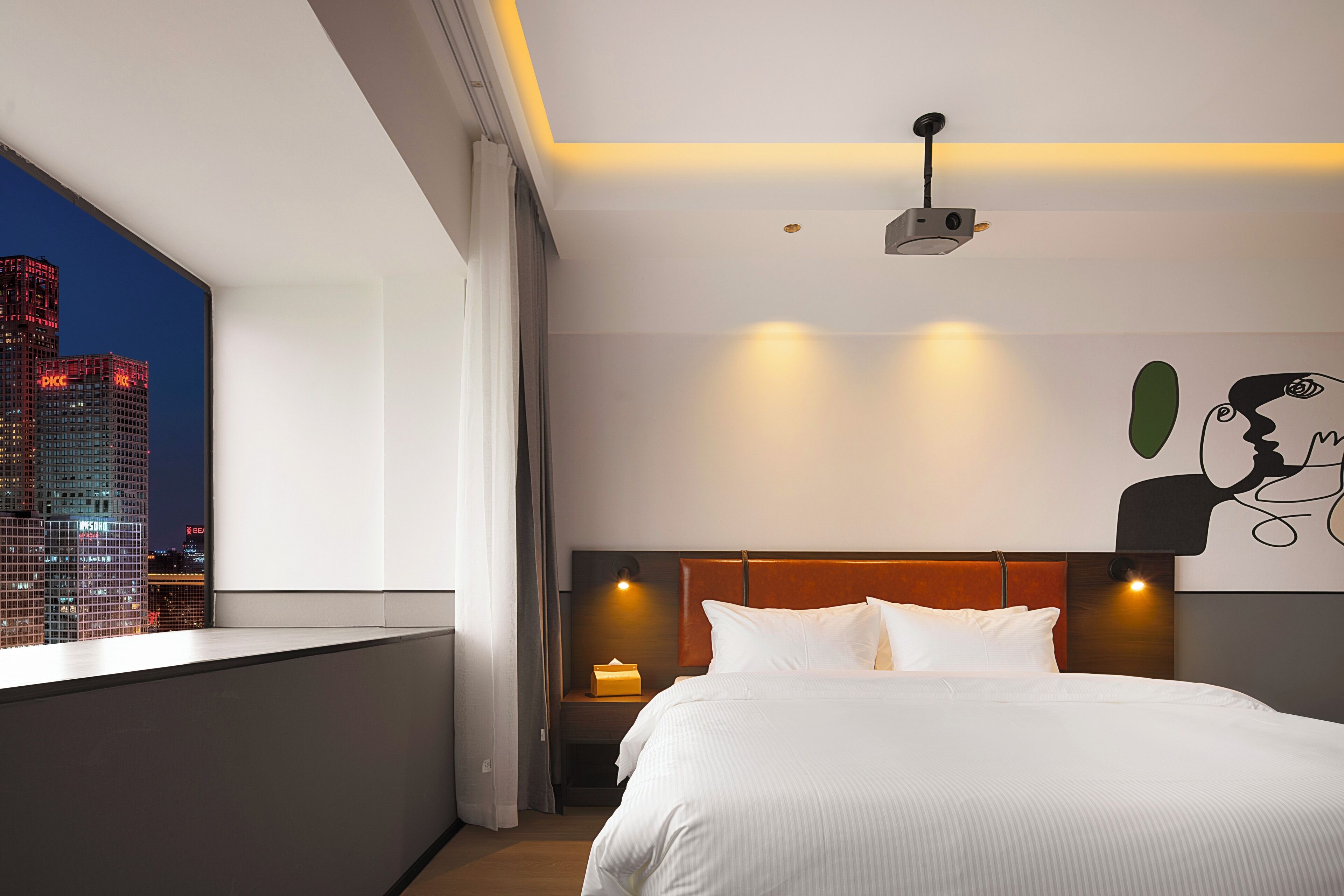Une chambre d’hôtel moderne dotée d’un grand lit, d’une table de chevet, d’un projecteur et offrant une vue sur la ville.