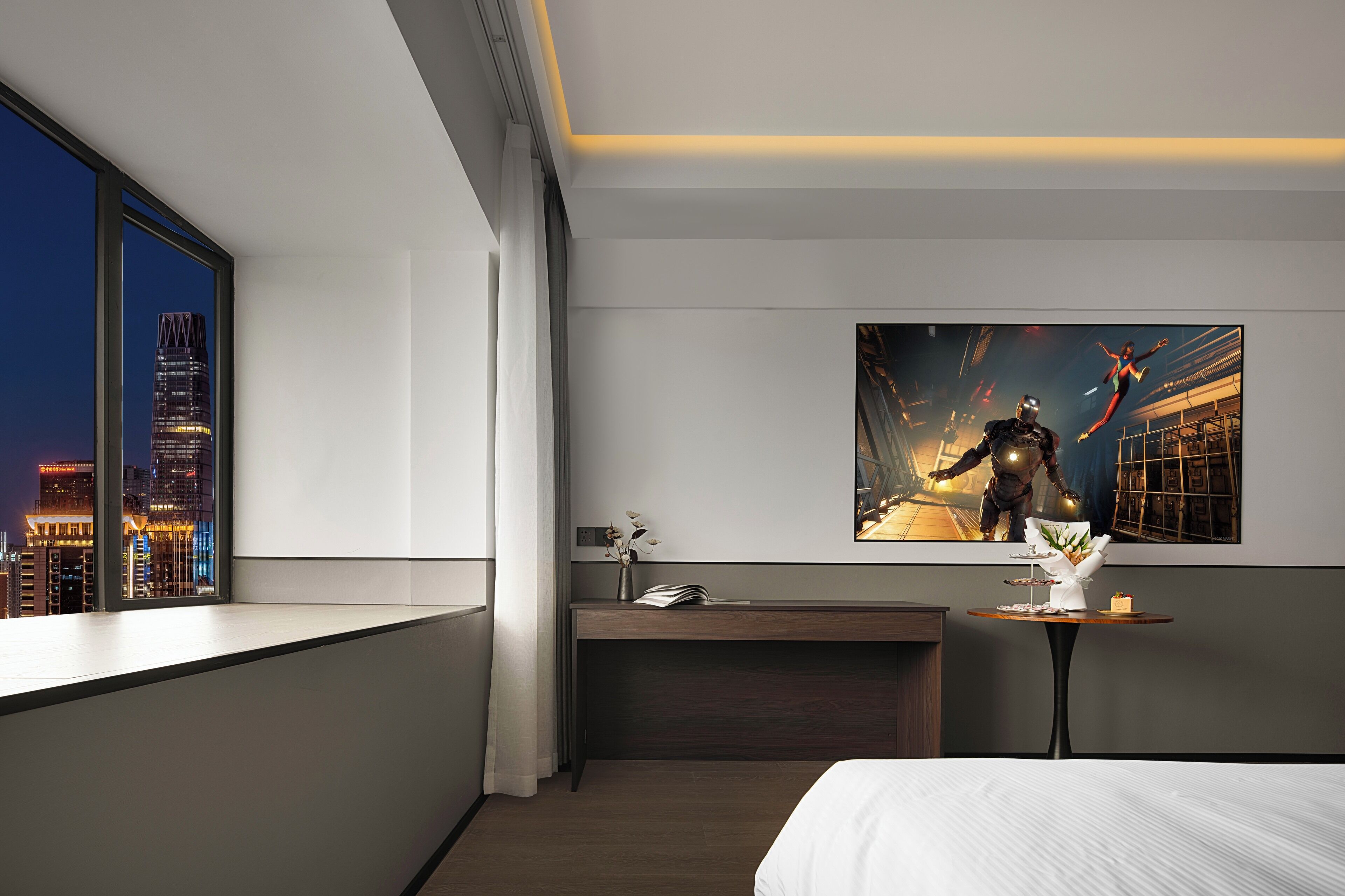 A modern hotel room with a city view, a desk with a vase of flowers, and a large poster on the wall.