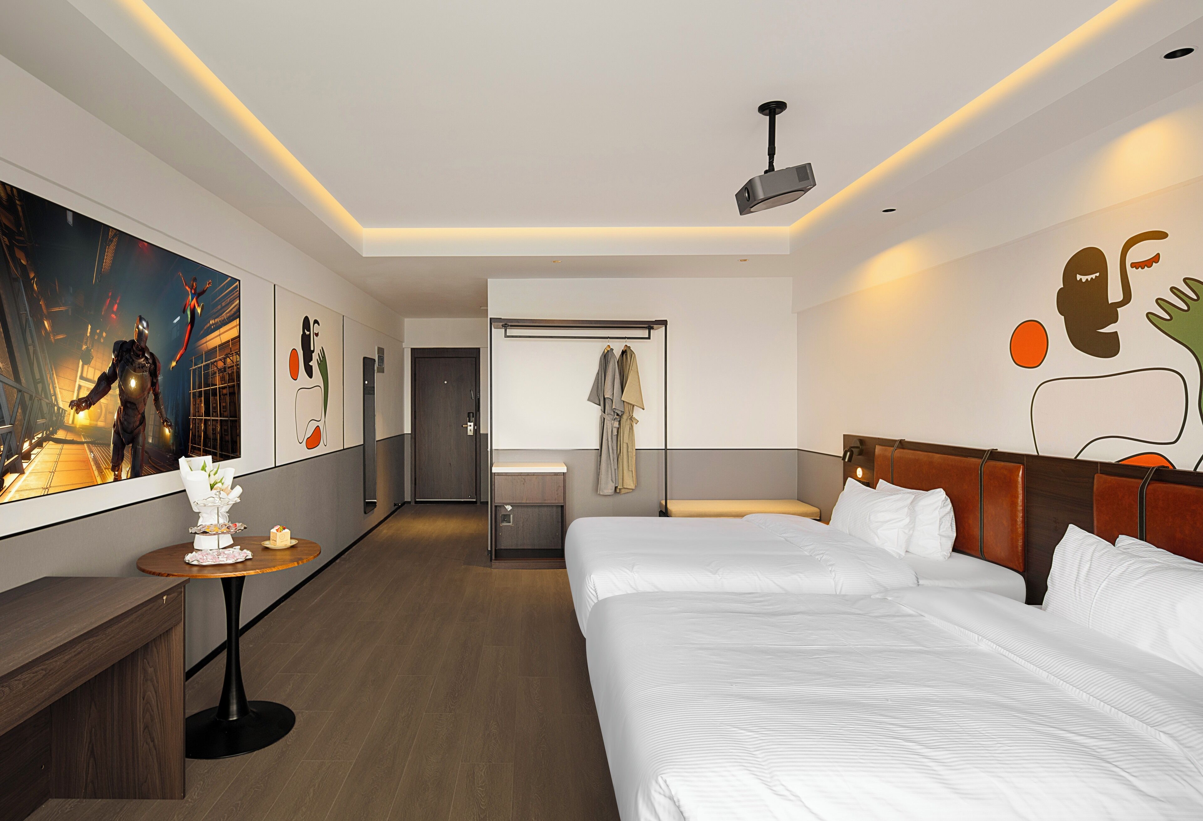 A hotel room with two beds, a small table with a vase and a cake, a coat rack, and a large painting on the wall.