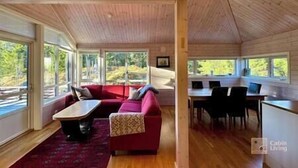 Ein Ferienhaus-Interieur mit einem roten Sofa, einem kleinen Tisch und einem Essbereich mit Stühlen. Große Fenster bieten Ausblicke ins Freie.