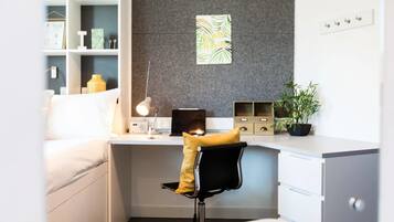 Un dormitorio moderno con cama blanca, un escritorio con una computadora portátil, una lámpara de escritorio y una pequeña planta en maceta.