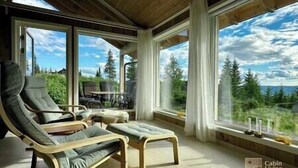 Une cabane avec deux fauteuils et une petite table, de grandes fenêtres offrant une vue sur les arbres et les montagnes, et un plafond en bois.