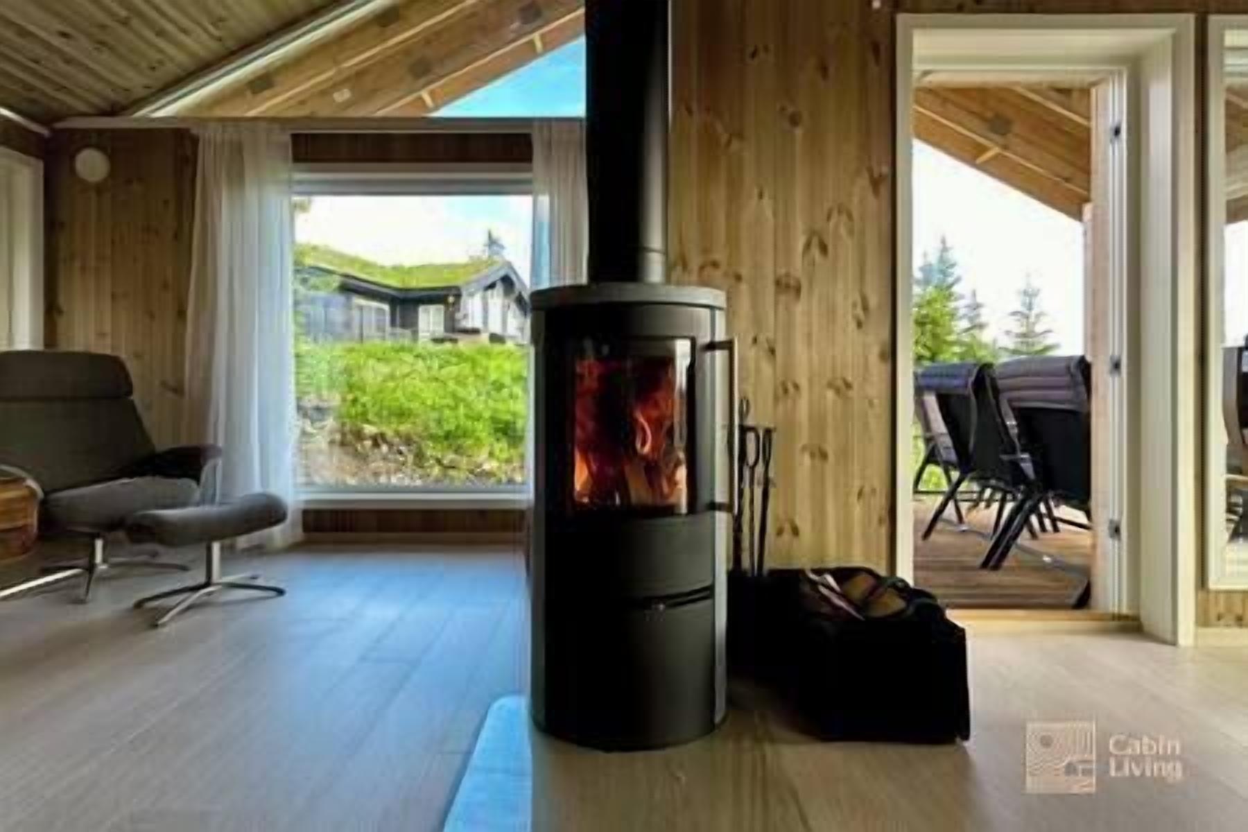 A modern cabin interior with a wood-burning stove, a comfortable chair, and a view of the outdoors.