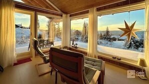 L’intérieur d’une cabane, avec de grandes fenêtres, un plafond en bois et une vue sur un paysage enneigé.