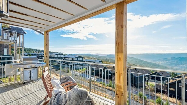 A wooden deck with a grill and seating, overlooking a mountainous landscape and modern buildings. - Ski in/out, bbq, mountain view & fireplace - Hafjell (Øyer kommune)