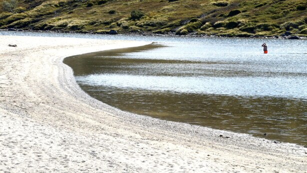 A beach with a curved shoreline, a person standing in the water, and a rocky hill in the background. - Cozy apartment in a central location in Sirdal (Sirdal kommune)