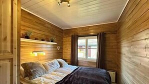 A wooden-paneled bedroom with a bed, a shelf with books, and a window with curtains. - Cozy cottage in Skeikampen - close to Hafjell! (Gausdal)