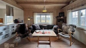 Interior de una cabaña con techo de madera, un sofá, un sillón mecededor, una mesa y una ventana con vistas.