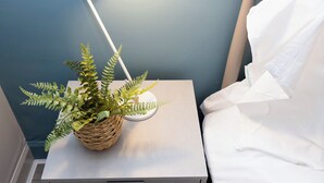 Ein Nachttisch mit einem gewebten Korb, der einen Farn, eine weiße Lampe und ein weißes Kopfkissen enthält.