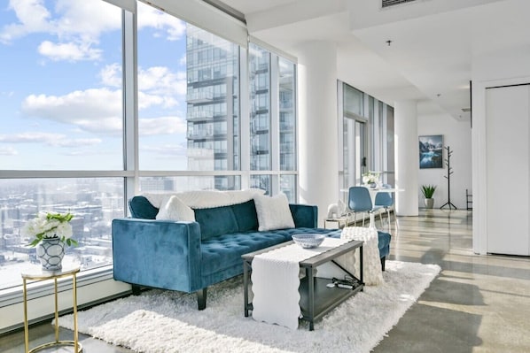 Ein modernes Wohnzimmer mit einem blauen Sofa, einem Couchtisch und Blick auf eine Stadtlandschaft durch große Fenster.