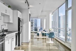 Eine moderne Küche mit Kühlschrank, Spüle und Schränken, die in einen Essbereich mit Tisch und Stühlen übergeht, und große Fenster mit Blick auf die Stadt.