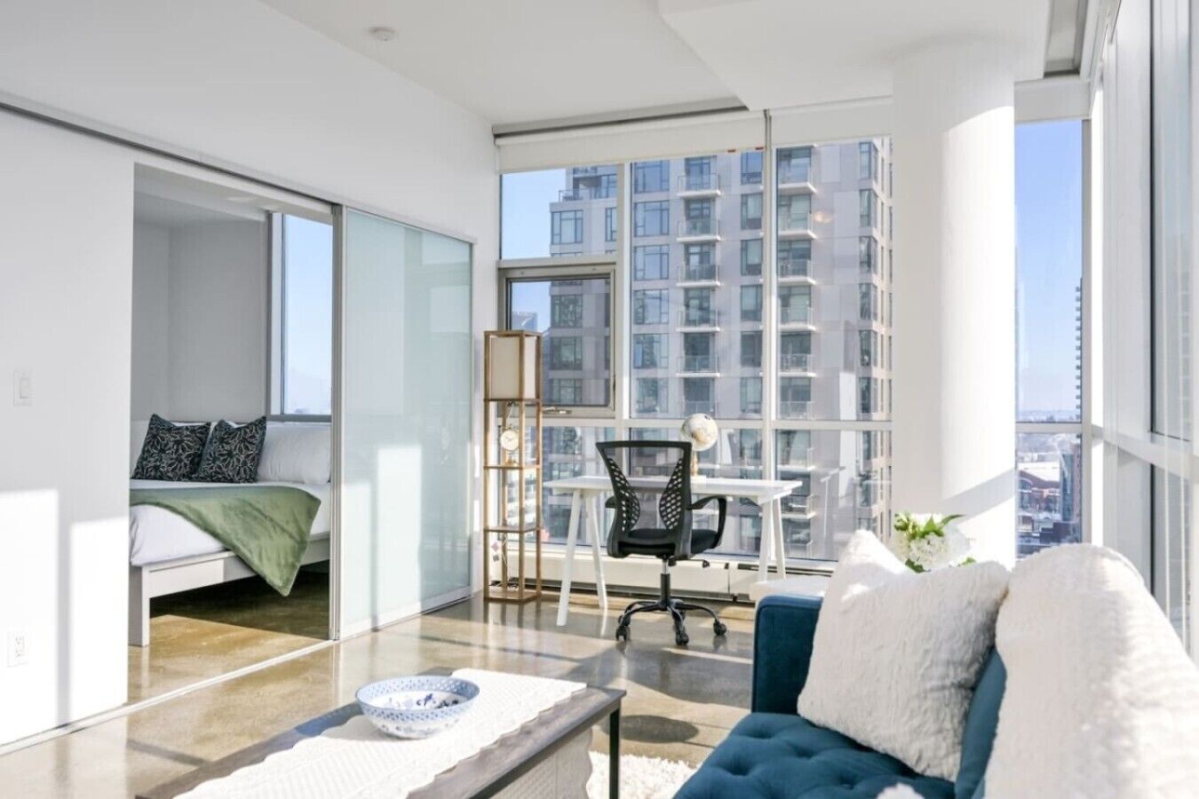 Un appartement moderne doté d’une grande fenêtre, d’un lit, d’un canapé bleu, d’une petite table et d’une chaise.