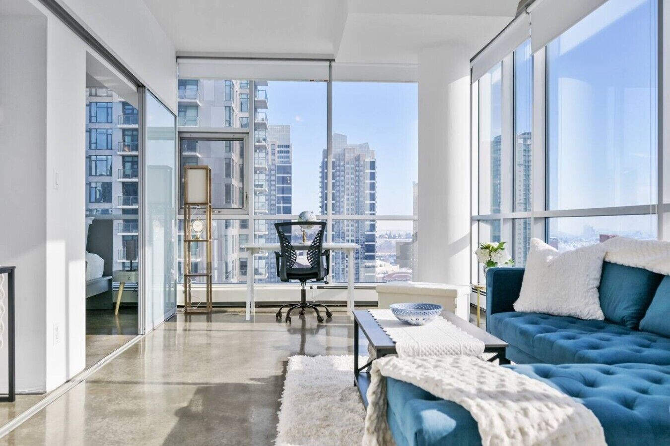 A modern living room with a sofa, a chair, a table, and a view of tall buildings through large windows.