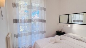 A bedroom with a bed, a nightstand, a lamp, a mirror, and a window with sheer curtains.