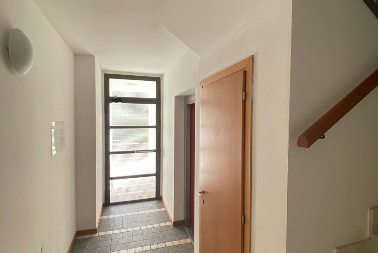 A narrow hallway with a wooden door on the right, a window with blinds, and a tiled floor.