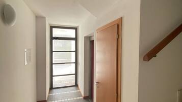 A narrow hallway with a wooden door on the right, a window with blinds, and a tiled floor.