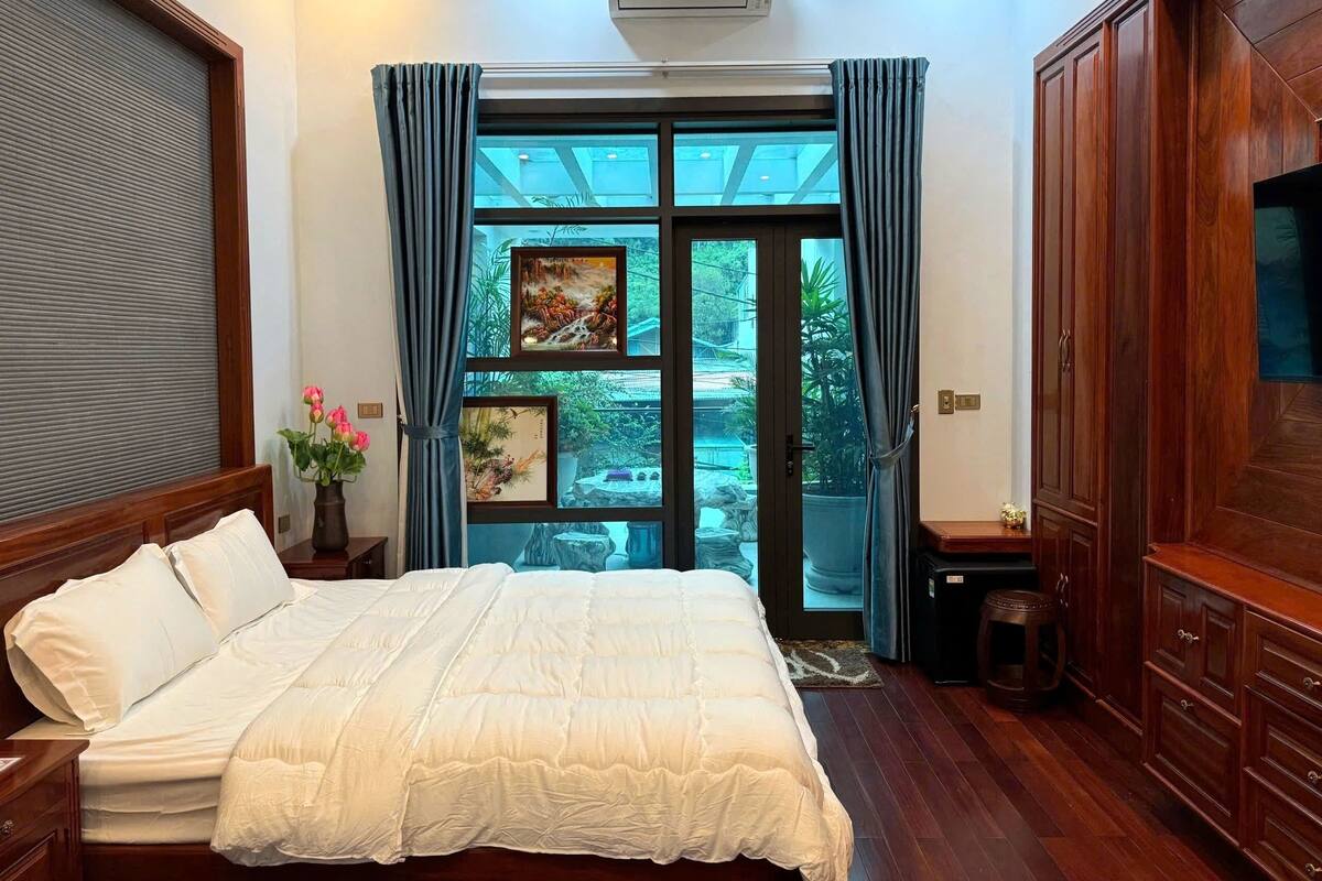 Un dormitorio con una cama grande, un armario de madera, un televisor y vistas a un jardín a través de un ventanal.