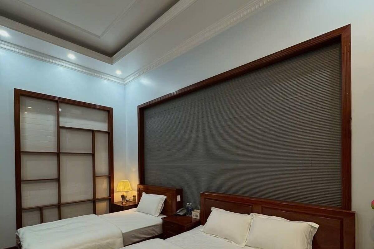 Habitación de hotel con dos camas, un ventanal con persianas, cabecera de madera y mesita de noche con lámpara.