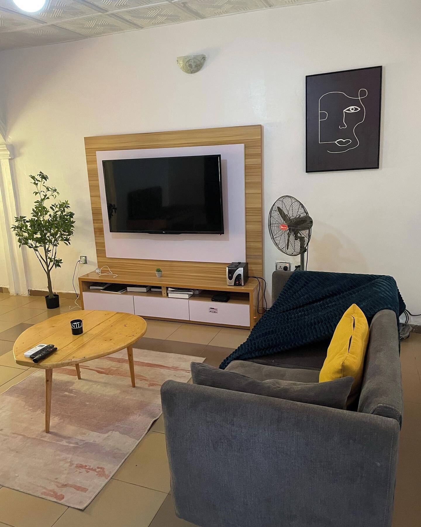 A living room with a large flat-screen TV, a wooden coffee table, a grey sofa, a small potted plant, and a wall-mounted fan.