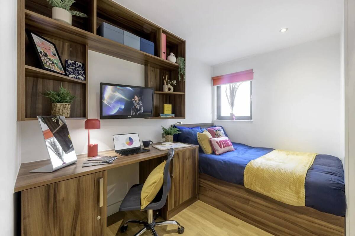 A bedroom with a wooden desk, a bed with a blue blanket, a TV, and shelves with storage boxes and decorative items.