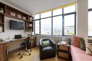 Enjoy a city view from the room's large window, featuring a desk, office chair, and cozy seating area with a sofa and decorative pillows.