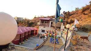 Un paysage rural avec un petit bâtiment, peut-être une maison d'hôtes, doté d'un toit rouge et d'un balcon. On peut voir des drapeaux colorés et un grand objet sphérique au premier plan.