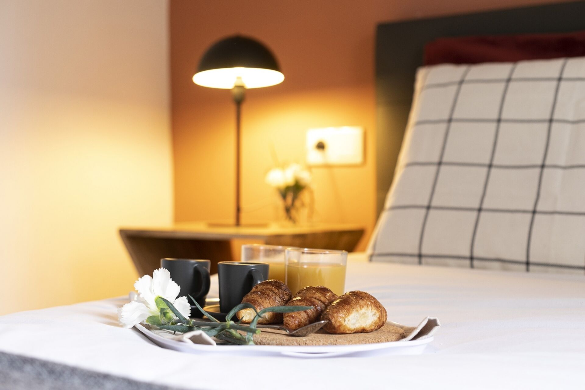 A breakfast tray with croissants, a flower, and two black cups on a bed with a bedside table, lamp, and a nightstand with a vase of flowers.