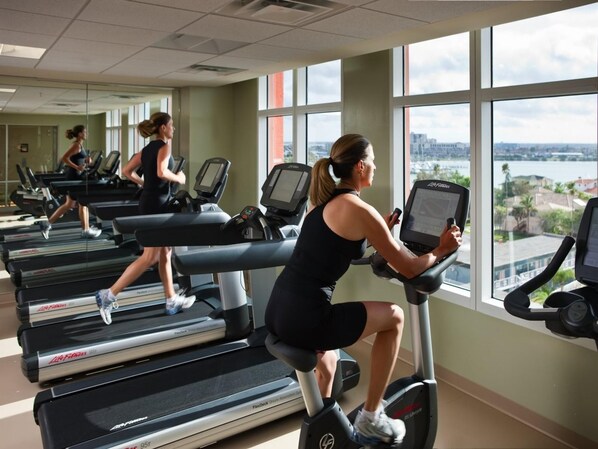 Un gimnasio con cintas de correr y bicicletas estáticas, amplias ventanas con vista a la ciudad y un espejo grande.