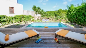 A wooden deck with two lounge chairs, a pool, and a tropical garden.