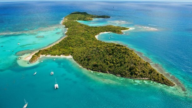 An aerial view of a tropical island with clear turquoise waters, surrounded by lush greenery and several boats anchored near the shore.