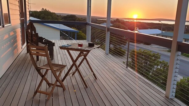 A wooden deck with a table and chairs, overlooking a sunset and a road with buildings in the distance.