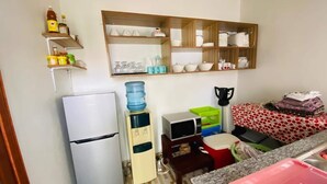 Eine kompakte Küche mit Kühlschrank, Mikrowelle und einer kleinen Arbeitsfläche. Darüber befinden sich Regale mit verschiedenen Küchenutensilien und ein Wasserspender.