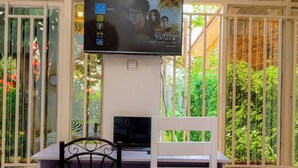 Ein Zimmer mit weißem Schreibtisch, zwei schwarzen Stühlen, einem an der Wand befestigten Fernseher und einem Fenster mit Blick auf Grünflächen.