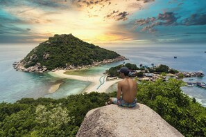 Una persona sentada sobre una roca, contemplando una playa con aguas cristalinas y una pequeña isla.