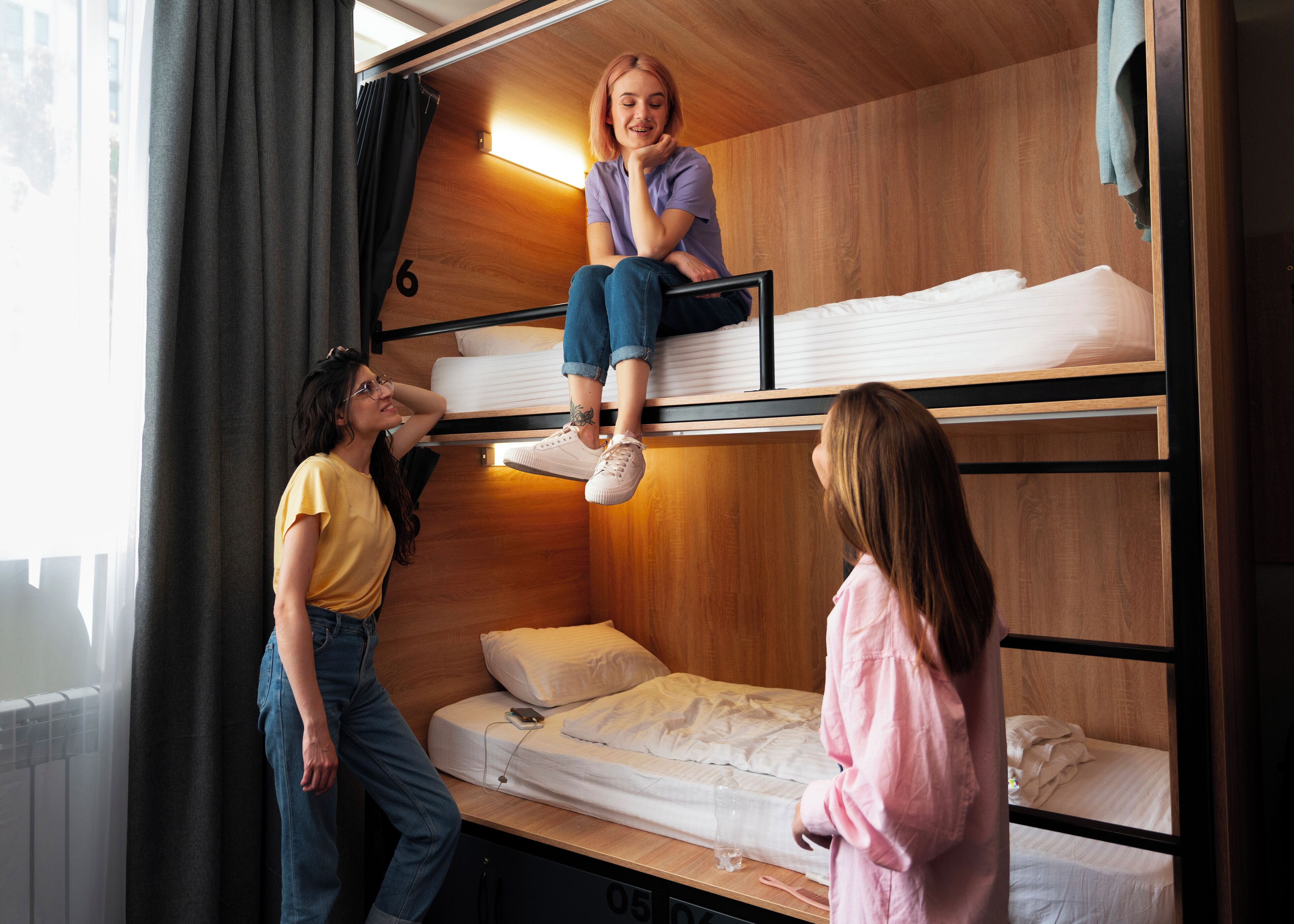 A bunk bed room with a person sitting on the top bunk, another person standing on the lower bunk, and a third person on the floor.
